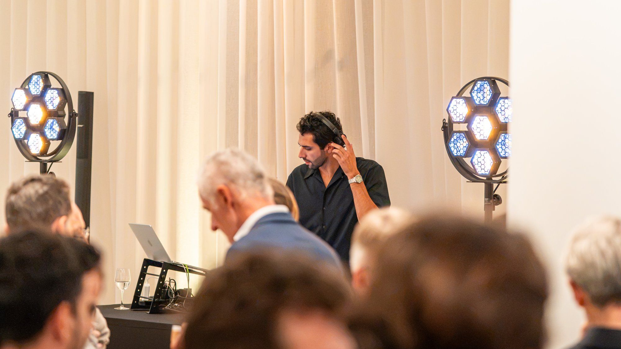 Le DJ, un casque sur l’oreille, derrière son ordinateur entre deux projecteurs, devant des invités au premier plan, lors d’un événement d’entreprise.