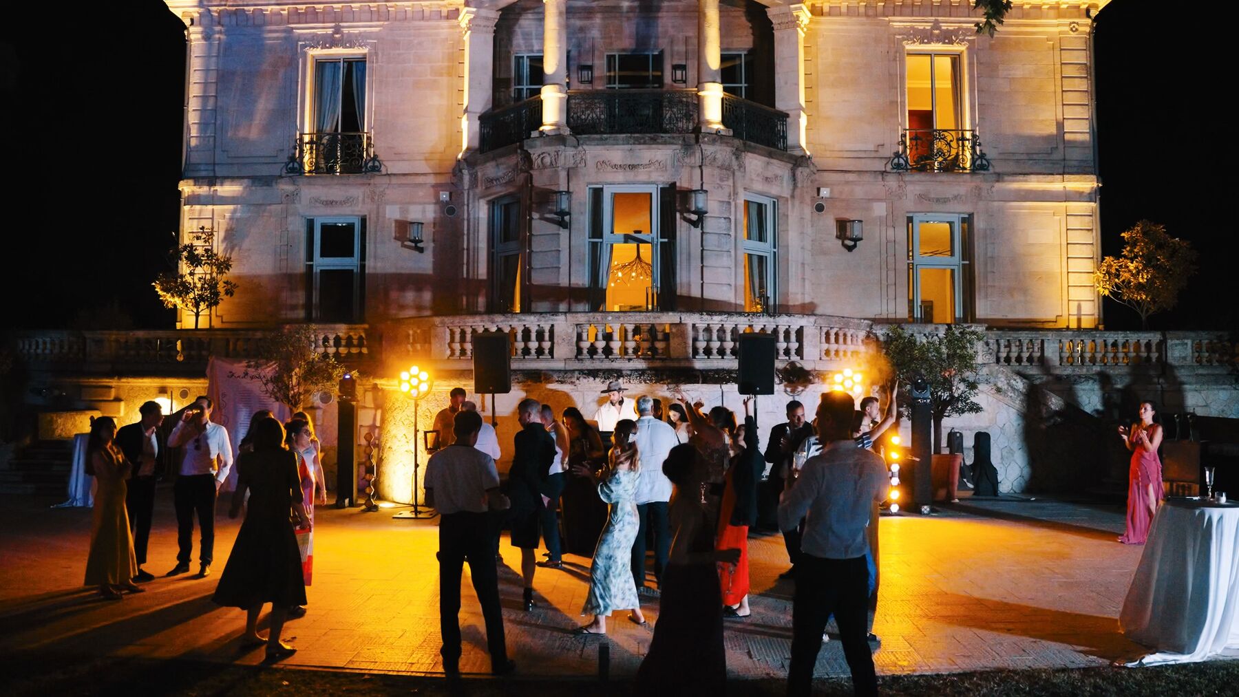An outdoor evening party in front of a lit-up château, with a DJ and guests dancing.