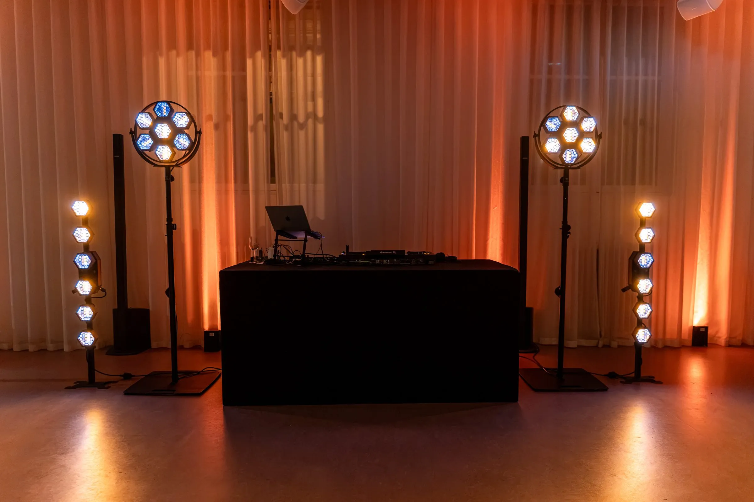 Installation son et lumière vue de face : console DJ sur une table noire, deux colonnes d’enceintes et quatre projecteurs à lentilles alvéolées devant un rideau orangé.