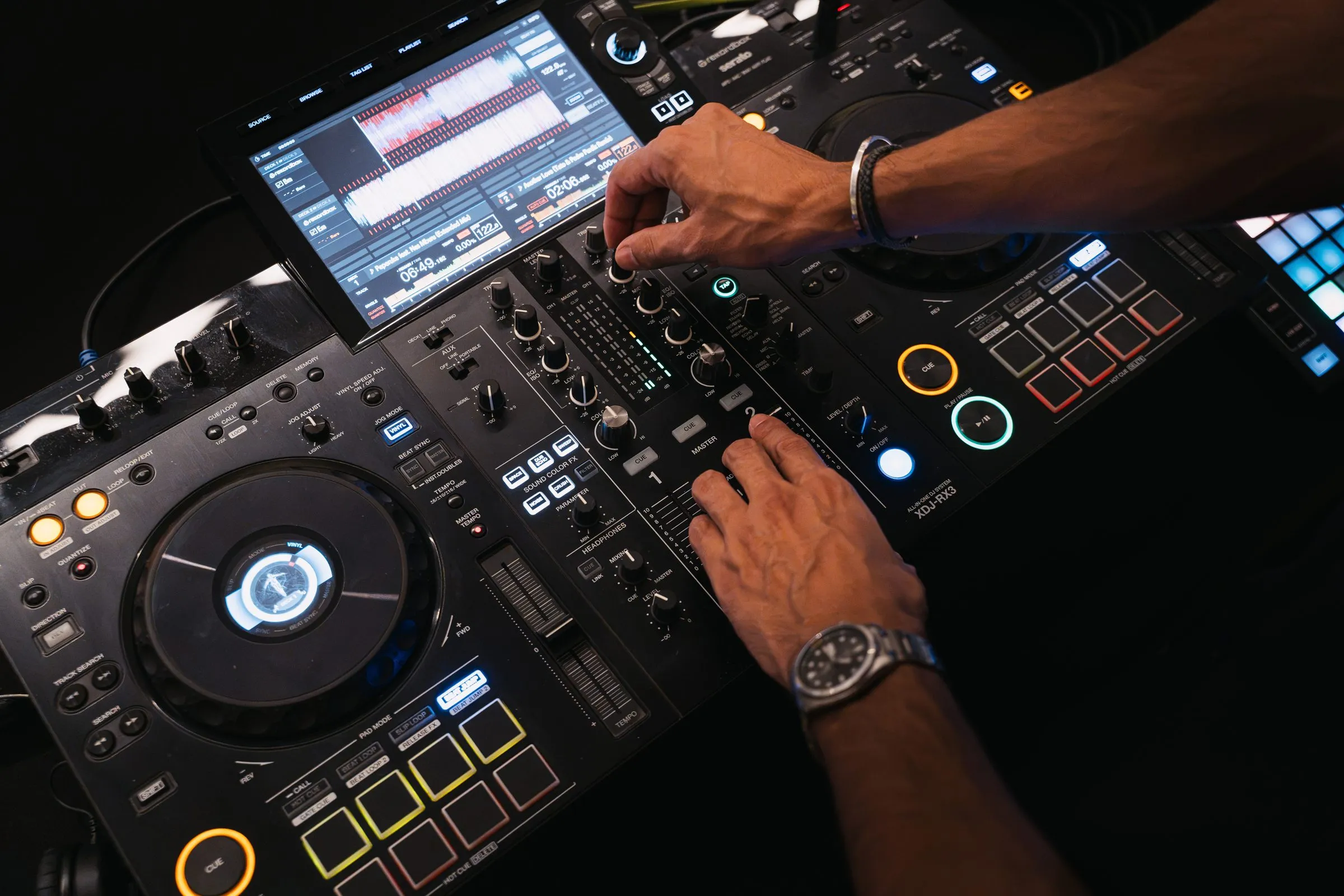 Vue plongeante sur des platines DJ : deux mains, une sur une molette et l’autre sur la table de mixage, sous un écran tactile.