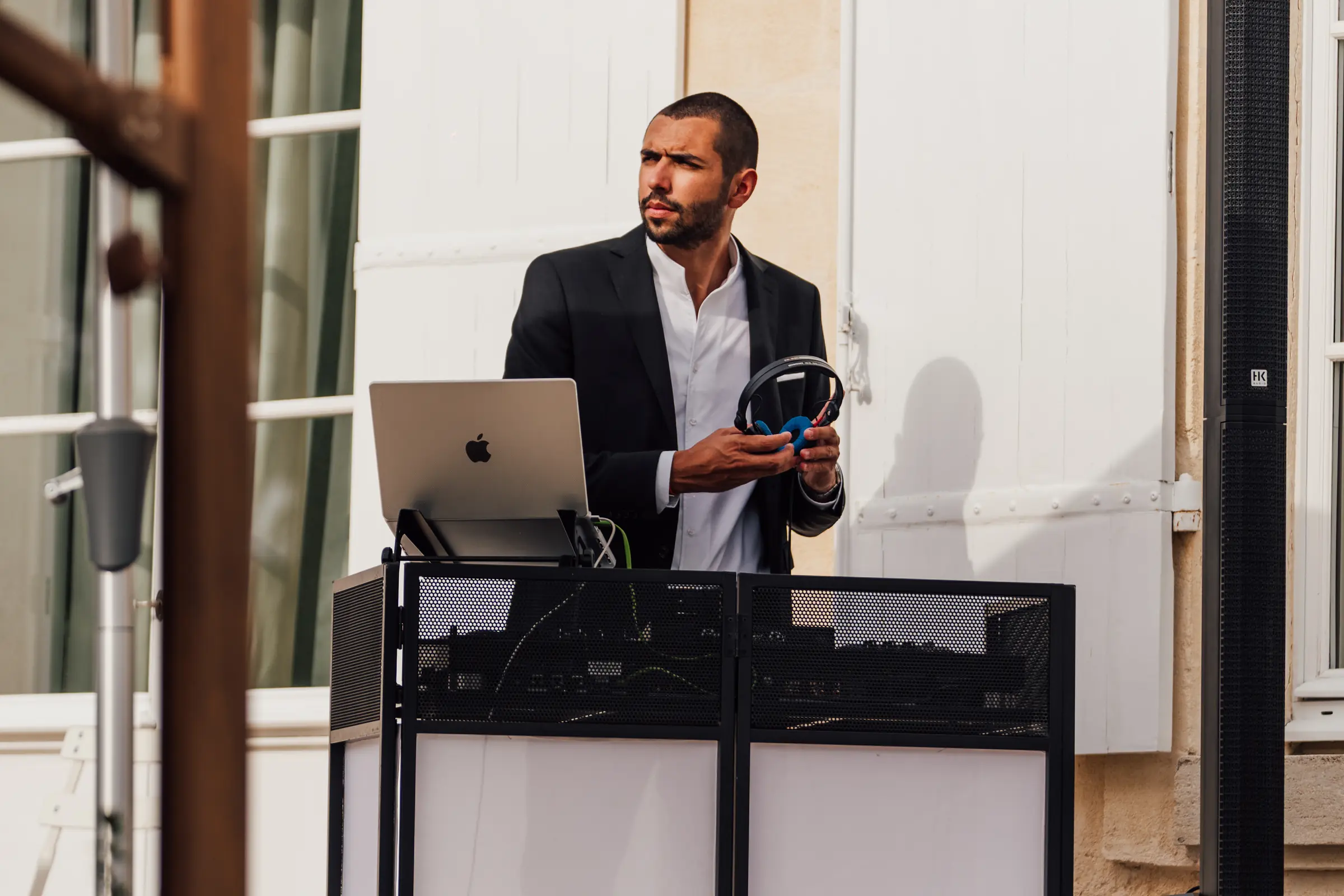 DJ en costume et chemise blanche derrière sa console blanche et noire, casque en main, sur une terrasse de château.