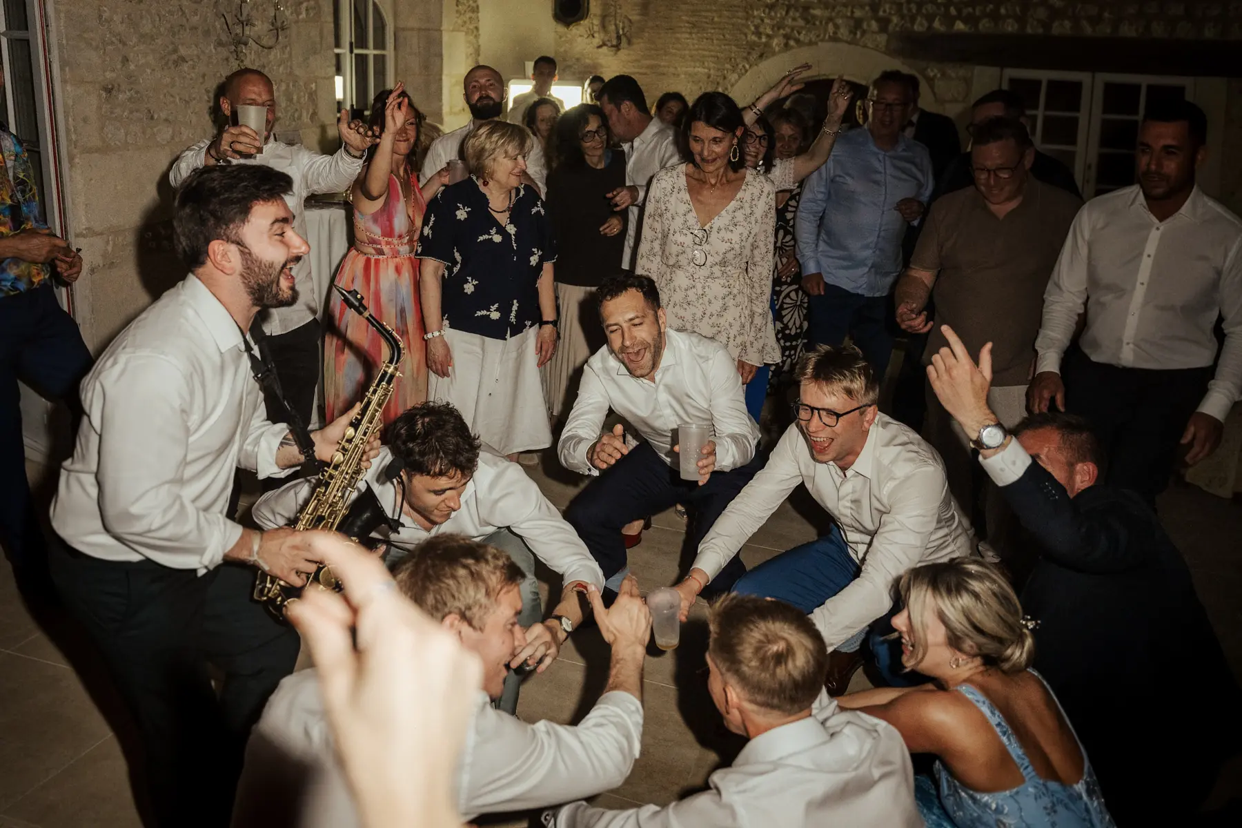 Un saxophoniste jouant au milieu des invités, en pleine soirée de mariage dans une salle en pierre.