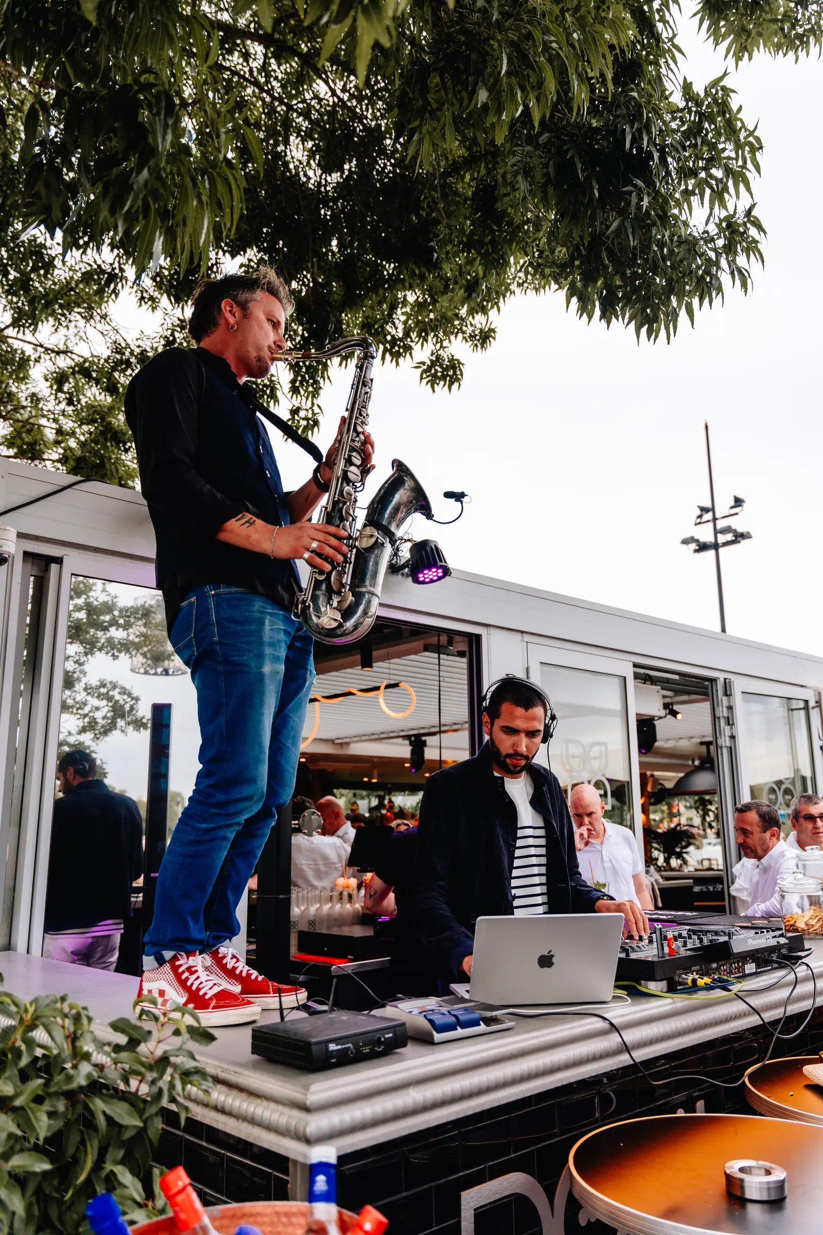 Saxophoniste jouant en plein jour sur une terrasse, DJ au casque en contrebas.