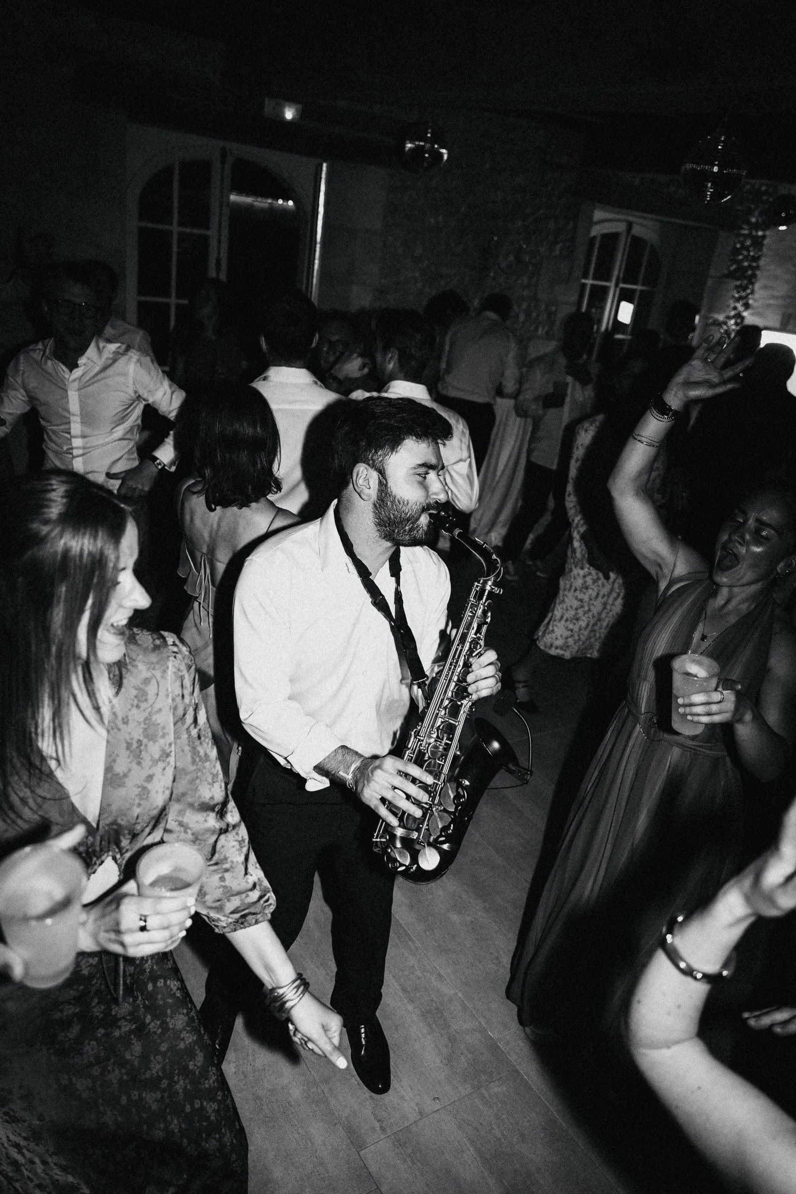 Un saxophoniste en chemise blanche joue au milieu des invités qui dansent, lors d’un mariage (noir et blanc).