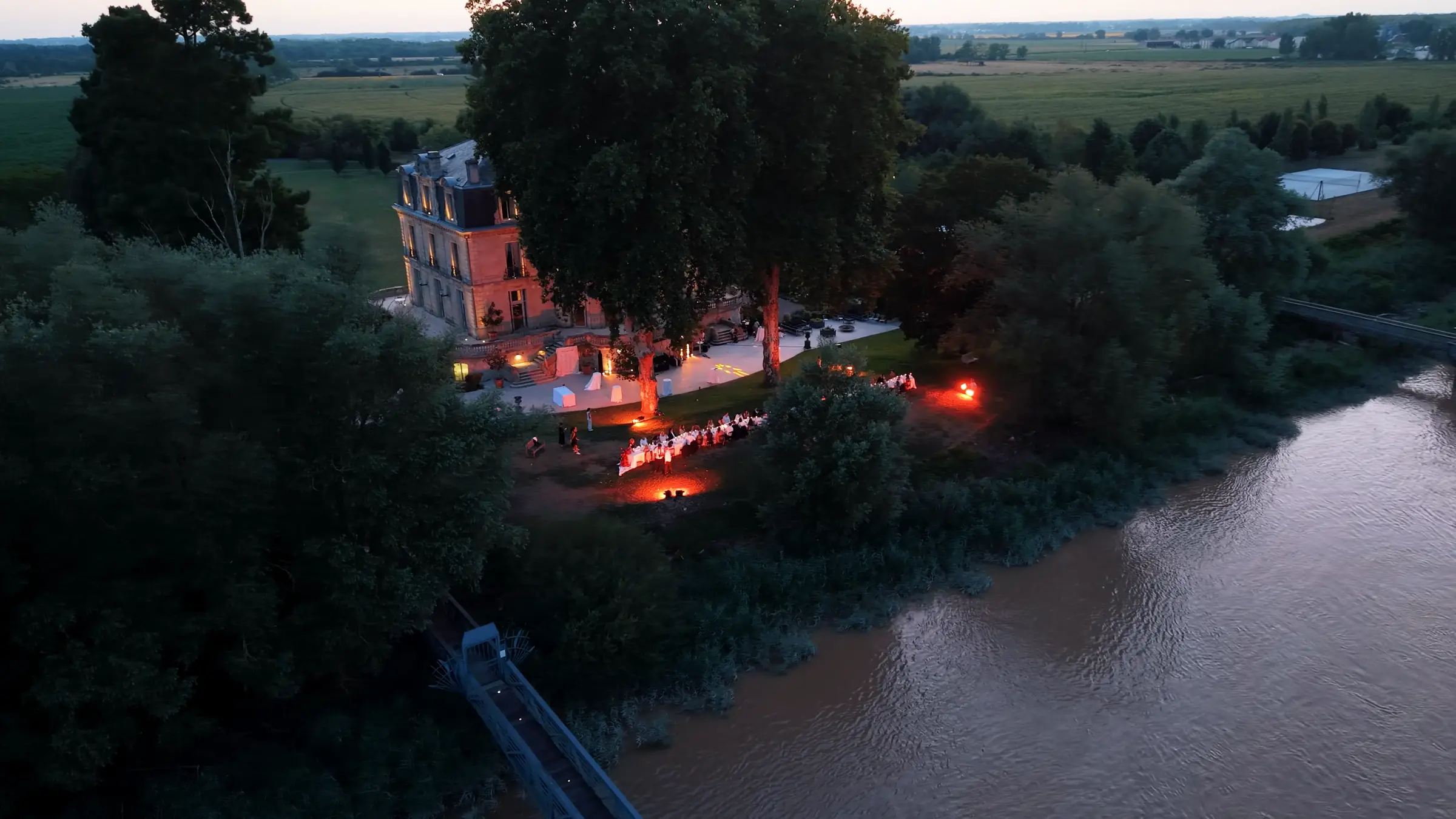 Vue aérienne d’un dîner de mariage éclairé par des flambeaux, au bord d’une rivière, château illuminé en arrière-plan, à la tombée de la nuit.